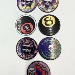 May include: Seven circular game tokens featuring various graphic designs. Designs include skulls with the text POISON, an eight ball with a skull and crossbones, a standard eight ball, and a skull surrounded by swirling colorful patterns.