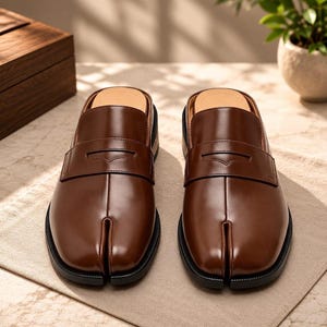 May include: A pair of brown leather slip-on loafers featuring a distinct split-toe design, a classic penny loafer strap across the vamp, and a smooth, polished finish. The shoes have a black sole and an open-back construction for easy wear.