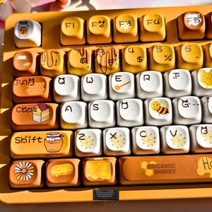 May include: A mechanical keyboard with a honey-themed design featuring yellow and white keycaps. The keys display illustrations of honey jars, bees, daisies, and honeycomb patterns, with the text "ORGANIC HONEY" printed on the spacebar.