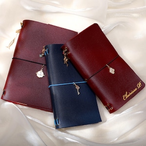 May include: Three leather journals in deep burgundy and navy blue. The journals feature elastic bands, metal charms including a key and fish, and gold-colored text on the rightmost cover that reads Clairissa P. The covers have visible eyelet details.