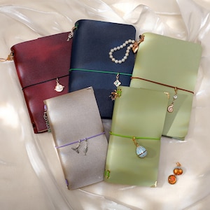 May include: Five leather journals in burgundy, navy blue, sage green, and metallic silver. Each notebook is secured with a colored elastic band and decorated with various metal charms, including a pearl strand, a shell, and a small locket.