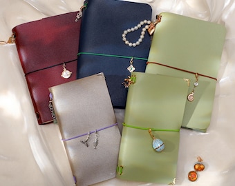 May include: Five leather journals in burgundy, navy blue, sage green, and metallic silver. Each notebook is secured with a colored elastic band and decorated with various metal charms, including a pearl strand, a shell, and a small locket.