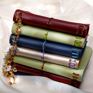 May include: A stack of six rectangular journals with smooth covers in shades of burgundy, light green, navy blue, and metallic silver. Each journal features metal eyelets with matching colored lacing and a gold-toned charm bracelet attached.