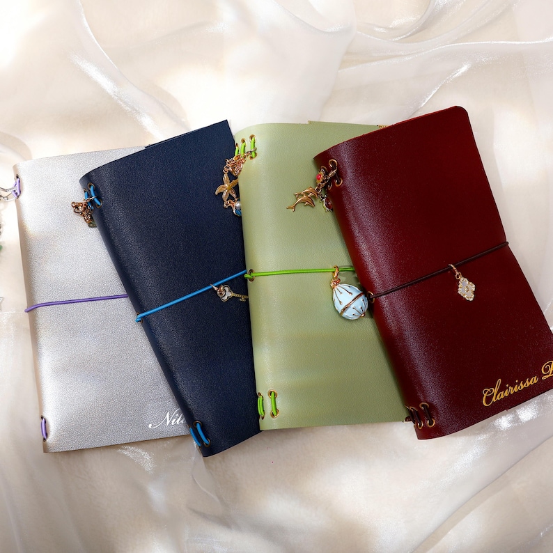 May include: Four rectangular leather journals in silver, navy blue, sage green, and deep burgundy. Each journal features a colored elastic closure band, metal eyelets, and decorative charms attached to the spine or elastic, with names embossed.