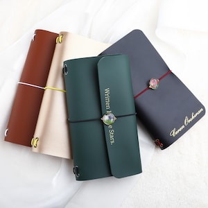May include: Four rectangular journals in brown, beige, dark green, and black. The green journal features the text Written In The Stars and a circular floral charm. The black journal has the name Ceren Öskıran and a floral charm on a red elastic band.