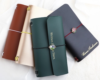 May include: Four rectangular journals in brown, beige, dark green, and black. The green journal features the text Written In The Stars and a circular floral charm. The black journal has the name Ceren Öskıran and a floral charm on a red elastic band.