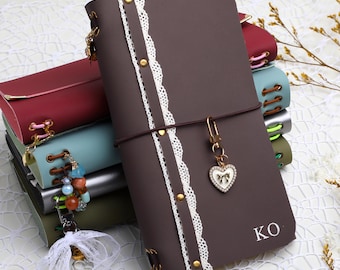May include: A dark brown leather-like journal featuring a vertical white lace strip secured with gold-tone rivets, a heart-shaped charm with a bow, and the letters KO printed in white. The journal is part of a stack of similar notebooks in various colors.