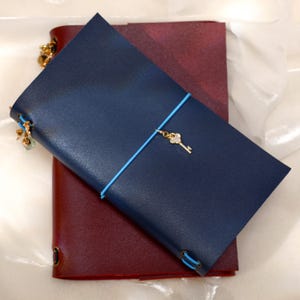May include: A dark blue leather journal cover with a light blue elastic band closure featuring a small gold-toned key charm. Beneath it lies a larger, deep burgundy leather journal cover. Both items feature visible elastic cord bindings.
