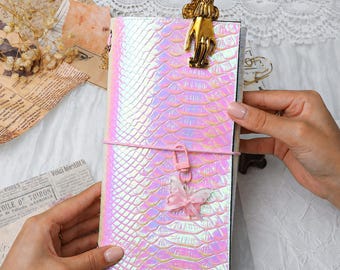 May include: A rectangular notebook with a holographic, iridescent snakeskin-textured cover. A gold-toned metal hand-shaped clip is attached to the top edge. A pink elastic band wraps around the center, featuring a small pink butterfly charm.