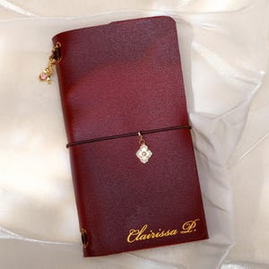May include: A deep burgundy leather journal cover featuring a gold elastic band closure, a gold clover-shaped charm, and a small pink dangling charm. The name Clairissa P. is embossed in gold script at the bottom right corner of the cover.