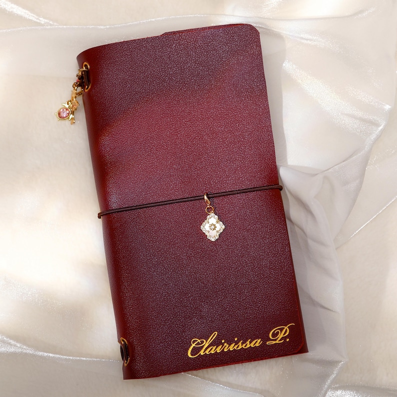 May include: A deep burgundy leather journal cover featuring a gold elastic band closure, a gold clover-shaped charm, and a small pink dangling charm. The name Clairissa P. is embossed in gold script at the bottom right corner of the cover.