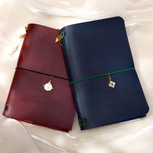 May include: Two leather-style journals, one in deep burgundy and one in navy blue, each secured with a colored elastic band. The burgundy journal features a gold-toned clock charm, while the navy journal has a square charm with a green center.