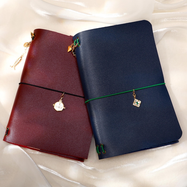 May include: Two leather-style journals, one in deep burgundy and one in navy blue, each secured with a colored elastic band. The burgundy journal features a gold-toned clock charm, while the navy journal has a square charm with a green center.
