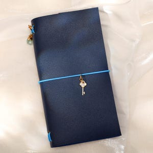 May include: A dark blue leather-textured notebook cover secured with a bright blue elastic band. A gold-toned key charm with a white inlay hangs from the center elastic. An additional small green bead charm is attached to the top left corner.