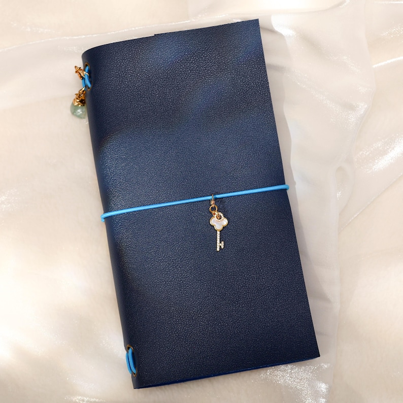 May include: A dark blue leather-textured notebook cover secured with a bright blue elastic band. A gold-toned key charm with a white inlay hangs from the center elastic. An additional small green bead charm is attached to the top left corner.