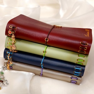 May include: A stack of four leather-like journals in burgundy, light green, navy blue, and metallic silver. Each journal features gold-toned metal eyelets with colored elastic cords and dangling charms attached to the side, with the name Clarissa D. embossed on the top cover.
