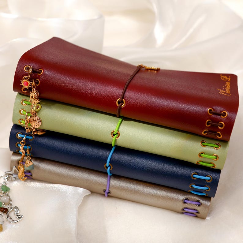 May include: A stack of four leather-like journals in burgundy, light green, navy blue, and metallic silver. Each journal features gold-toned metal eyelets with colored elastic cords and dangling charms attached to the side, with the name Clarissa D. embossed on the top cover.
