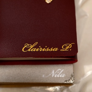 May include: A deep burgundy notebook with the name Clairissa P. embossed in gold script on the cover, stacked atop a light gray notebook featuring the name Nila in white script and a decorative silver metal corner accent on the bottom right.
