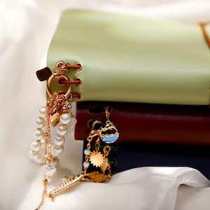 May include: A stack of three leather-bound journals in light green, dark red, and navy blue. Each journal features gold-toned metal eyelets with brown cord lacing and decorative gold-toned charms, including pearl beads, a dolphin, and a fish.