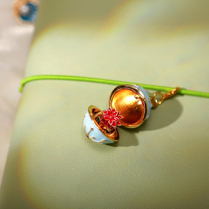 May include: A gold-toned metal locket charm shaped like a sphere with a light blue exterior and a bright red flower inside. The locket is open, revealing the flower, and is attached to a thin, bright green cord that runs horizontally across the frame.