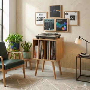Puede incluir: Un mueble para tocadiscos de madera clara con cuatro patas cónicas y tres compartimentos verticales de almacenamiento llenos de discos de vinilo. Un tocadiscos negro descansa encima y cinco portadas de álbumes enmarcadas cuelgan.
