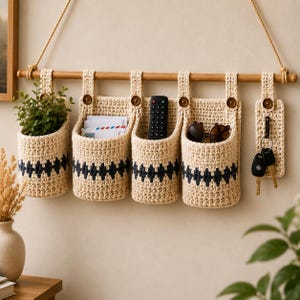 Op de afbeelding: Een wandorganizer met vier crèmekleurige gehaakte zakken met donkergrijze geometrische patronen, opgehangen aan een houten stok met een touw. De zakken bevatten een kleine groene plant, post, een afstandsbediening, een zonnebril en een sleutelhaak.