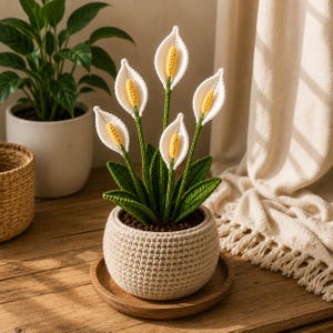 May include: A crocheted peace lily plant featuring five white spathes with yellow spadices, green leaves, and a textured beige crocheted pot. The plant is arranged in a round, neutral-toned container that sits on a circular wooden base.