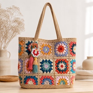Może przedstawiać: Torba typu tote wykonana z beżowej włóczki z wielokolorowym wzorem szydełkowym typu granny square. Torba posiada dwa paski na ramię oraz ozdobną zawieszkę składającą się z szydełkowanego kwiatka i dwóch chwostów w kolorze różowym i morskim.