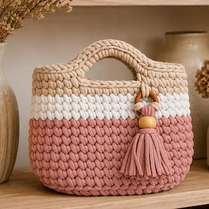 Puede incluir: Un bolso de ganchillo con un diseño de bloques de color con rayas horizontales en beige, blanco y rosa empolvado. El bolso tiene asas integradas y está decorado con una borla de tela a juego sujeta a una anilla y una cuenta de madera.