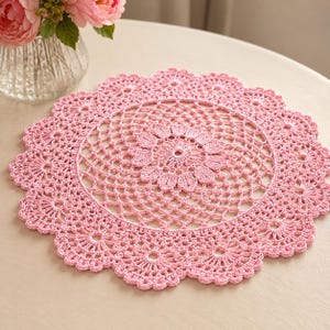 Puede incluir: Un tapete de ganchillo redondo de color rosa claro que presenta un intrincado patrón floral calado en el centro, rodeado por un delicado diseño de celosía y un borde festoneado en forma de concha. La pieza textil está hecha con hilo fino.