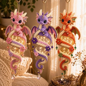 May include: Three hanging crochet dragon ornaments in pink, purple, and orange. Each dragon features large black eyes, wings, and a coiled body wrapped around a glowing, spherical light. The designs are accented with small flowers and beads.