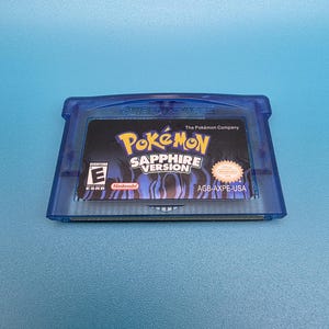 May include: A translucent blue Game Boy Advance video game cartridge for Pokémon Sapphire Version. The label features the game title, the ESRB Everyone rating, the Nintendo logo, and the product code AGB-AXPE-USA on a dark background.