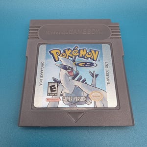 May include: A grey Nintendo Game Boy video game cartridge for Pokémon Silver Version. The label features the legendary Pokémon Lugia, the Pokémon GS logo, the ESRB Everyone rating, and the text DMG-AAXE-USA and THIS SIDE OUT.
