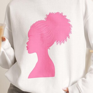 May include: A white long-sleeve crewneck sweatshirt featuring a bright pink graphic print of a side profile silhouette of a person with a large curly bun hairstyle. The sweatshirt is paired with gray denim shorts and a clear plastic cup with a straw.