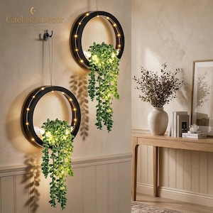 May include: Two circular black wall-mounted planters featuring integrated warm LED string lights. Each ring holds a trailing green artificial succulent plant with small, rounded leaves. The planters are suspended by thin wires from black hooks.