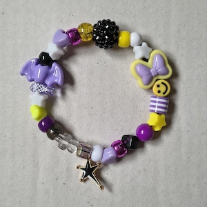 Könnte beinhalten: Ein Perlenarmband mit einer Vielzahl bunter Kunststoffperlen in Lila, Gelb, Schwarz und Weiß. Das Design umfasst geformte Perlen wie eine lila Fledermaus, einen gelben Schmetterling, ein gelbes Smiley-Gesicht und einen hängenden Sternanhänger.