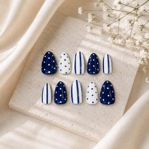 May include: A set of ten almond-shaped press-on nails featuring a nautical color palette of navy blue and white. The design alternates between solid navy blue with white polka dots, solid white with navy blue polka dots, and vertical navy blue stripes.
