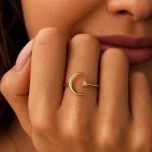 May include: A gold-toned open ring featuring a crescent moon motif on one end and a small, five-pointed star on the other. The star is accented with a single, clear, round-cut gemstone set in the centre, creating a celestial-inspired design.