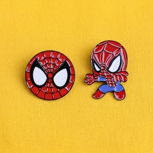 May include: Two enamel pins featuring a superhero character. One pin is a circular red mask with a black web pattern and large white eyes. The second pin depicts the full character in a red and blue suit with a black web design and white eyes.