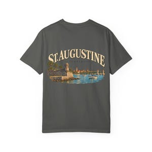 Può includere: Una t-shirt grigio antracite a maniche corte con una grafica sul retro. Il disegno raffigura una scena costiera con un forte in pietra, barche a vela e un ponte, con il testo ST. AUGUSTINE FLORIDA in lettere color crema arcuate sopra l'immagine.