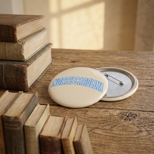 May include: A round pinback button with a cream-colored background and the text NORTH CAROLINA printed in a light blue and white checkered pattern. The button features a metal back with a pin mechanism for attachment to clothing or fabric items.
