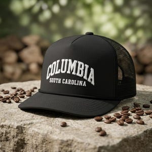May include: A black trucker hat featuring a mesh back and a solid front panel. The front displays white embroidered text that reads COLUMBIA in a large arched font, with SOUTH CAROLINA printed in a smaller, straight font directly underneath.