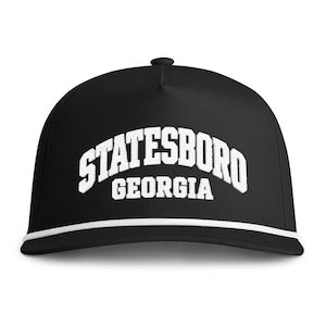 May include: A black baseball cap featuring white embroidered text that reads STATESBORO GEORGIA in an arched collegiate font. The cap includes a white braided rope detail across the base of the crown, just above the flat brim, and a top button.