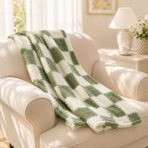 Puede incluir: Una manta de felpa con un patrón de cuadros repetitivo en cuadrados alternos de color verde salvia y blanco crema. La tela tiene un pelo grueso, difuso y texturizado, drapeado sobre el asiento y el respaldo de un sillón de color neutro.