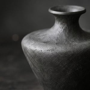 以下が含まれることがあります： マットブラックのセラミック製花瓶。幅広で丸みを帯びた胴体と、縁が広がった細い首が特徴です。表面全体には、細い線が多方向に刻まれた緻密なテクスチャーが施されており、触感のある仕上がりになっています。