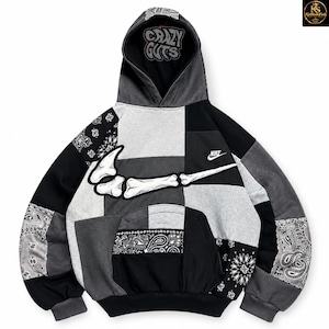 May include: A black, gray, and white patchwork hoodie featuring a large white bone-shaped Nike swoosh graphic on the chest. The interior hood lining displays the text "CRAZY GUTS" in a stylized font. The garment includes black and white paisley patterns.