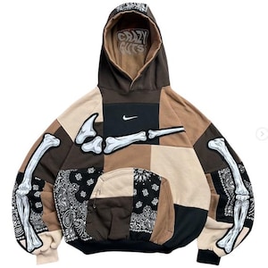 May include: A patchwork hoodie in shades of brown, tan, and black with a white Nike swoosh logo. The garment features a white bone graphic across the chest and sleeves, black bandana-patterned fabric panels, a front pocket, and the text Crazy Guts.