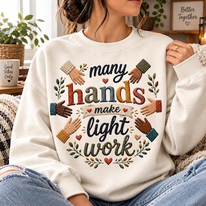 Puede incluir: Una sudadera de cuello redondo color crema con un estampado gráfico colorido de varias manos dispuestas en círculo alrededor del texto "many hands make light work" en azul oscuro, rojo y dorado, acentuado con pequeños corazones rojos y hojas.