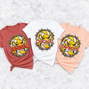 Op de afbeelding: Drie t-shirts in baksteenrood, wit en licht perzik. Elk shirt heeft een afbeelding van een geel-witte volleybal met een boos gezicht, omringd door prikkeldraad. De shirts tonen de teksten ANNA, CHICAGO en COWBOYS op de afbeeldingen.