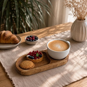 May include: A wooden serving tray holds a white ceramic mug filled with latte, a small fruit tart topped with red berries and blueberries, and a round cookie. Nearby, a plate features a golden-brown croissant, and a small bowl contains mixed berries.